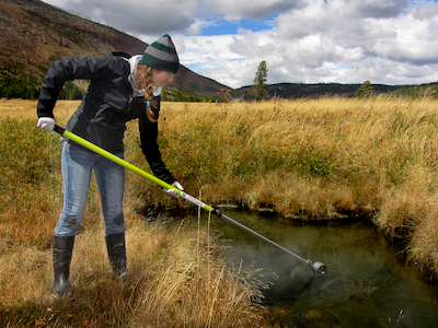 Student using pole to sample water from water body in Yellowstone Park, mountains in background