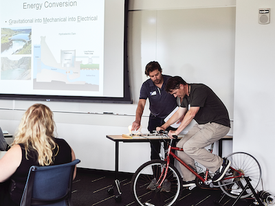 professor stands by k-12 teacher pedalling stationary bicycle as demonstration of generating electricity